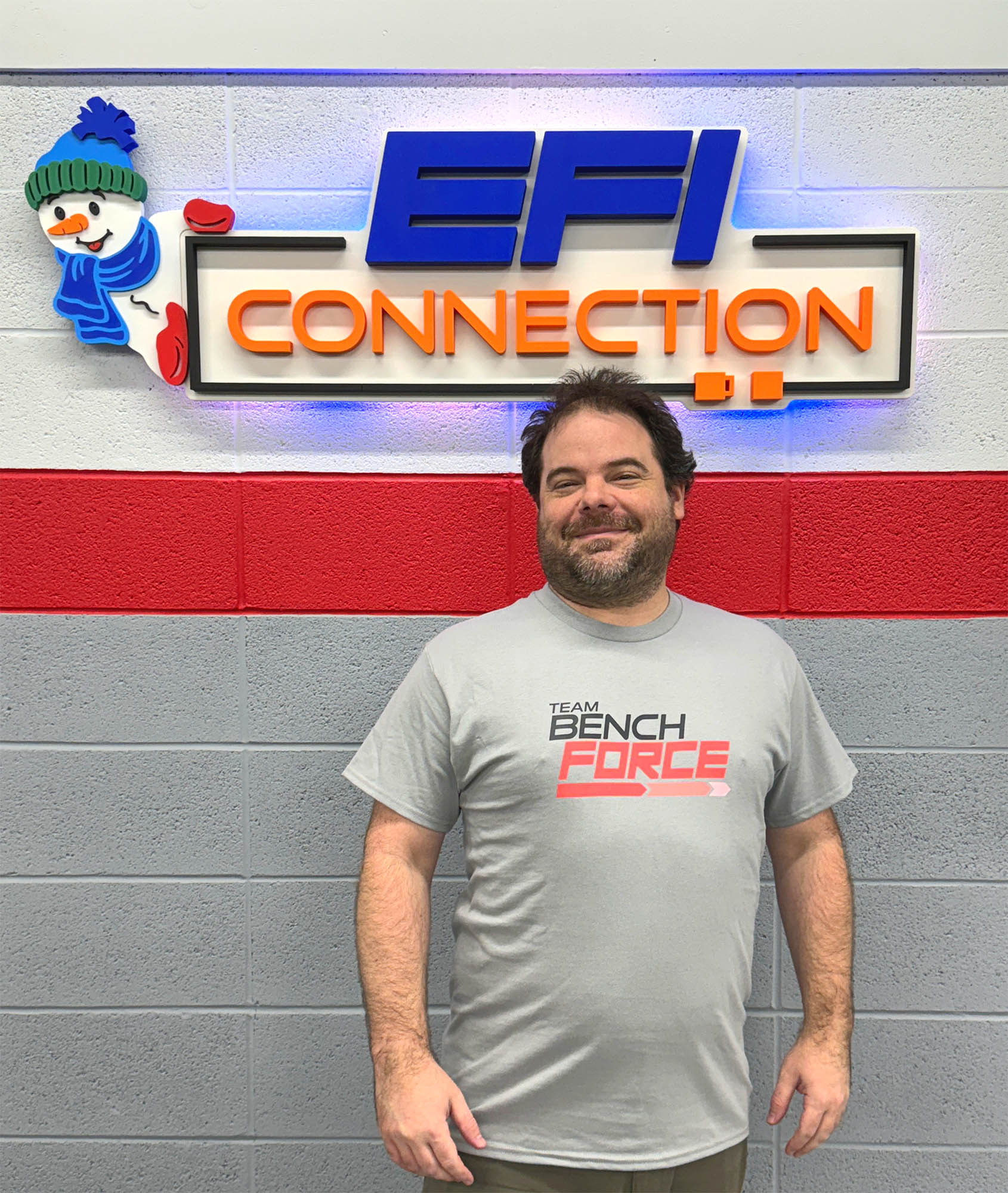A man standing in front of a sign that reads 'EFI CONNECTION', wearing a gray t-shirt with 'BENCHFORCE' text on it.